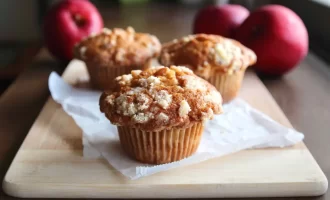 Three apple crumble muffins sit on a wooden board with parchment paper. In the background are three ripe red apples.