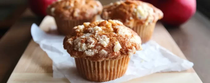 Three apple crumble muffins sit on a wooden board with parchment paper. In the background are three ripe red apples.
