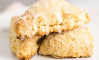 Close-up of two golden-brown scones stacked on a white plate, drizzled with glaze. The scones appear fresh and crumbly.