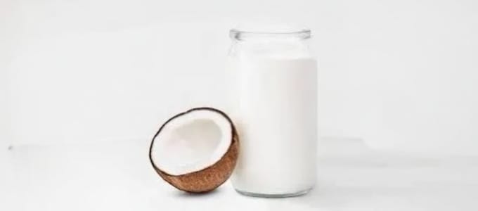 A glass jar of white coconut milk stands beside a halved coconut on a plain white surface. The image conveys a fresh and natural feel.