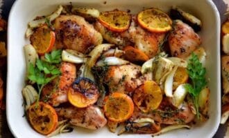 Roasted chicken thighs in a baking dish with sliced oranges, fennel, and herbs. The dish appears aromatic and delicious, with a garnish of fresh parsley.