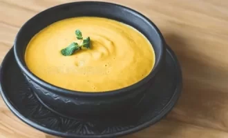 A black bowl of creamy pumpkin or squash soup garnished with fresh herbs sits on a matching black plate on a wooden.