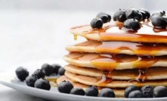 A stack of pancakes topped with fresh blueberries and drizzled with maple syrup on a white plate. Light gray background.