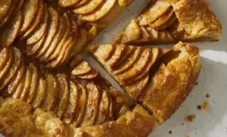 A golden-brown apple galette on a white surface, featuring neatly arranged apple slices forming a spiral pattern. Two triangular slices are cut, revealing a flaky crust.