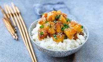 A bowl of white rice topped with glazed chicken pieces and green onions, sprinkled with sesame seeds.