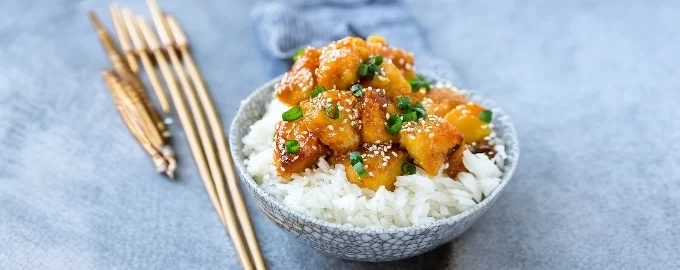 A bowl of white rice topped with glazed chicken pieces and green onions, sprinkled with sesame seeds.