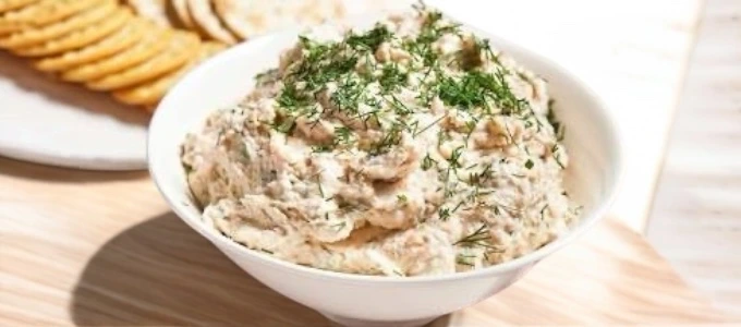 Creamy dip topped with fresh dill in a white bowl, set on a wooden table. A plate of sliced crackers is in the background, suggesting a snack setting.