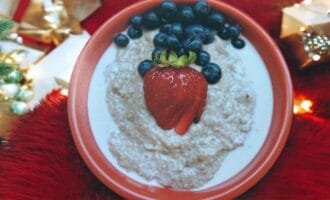 A festive breakfast bowl featuring oatmeal topped with a strawberry and blueberries is surrounded by Christmas.