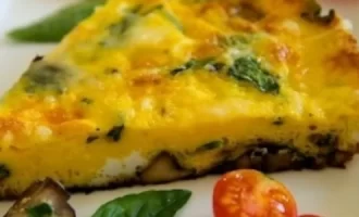 A slice of frittata with spinach and mushrooms on a white plate, garnished with cherry tomatoes and a fresh basil leaf.