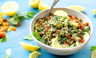 A tabbouleh bowl of couscous salad with vegetables and herbs sits on a light blue table. Fresh lemon wedges.