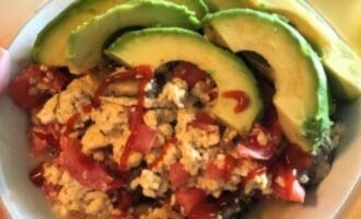 A bowl of scrambled eggs mixed with diced tomatoes, topped with avocado slices