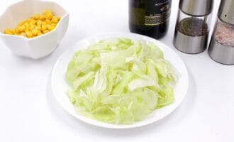 Plate of sliced lettuce on a white surface, accompanied by a bowl of corn, a bottle of olive oil, and salt