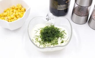 A glass bowl of creamy sauce topped with fresh dill alongside a dish of corn kernels.